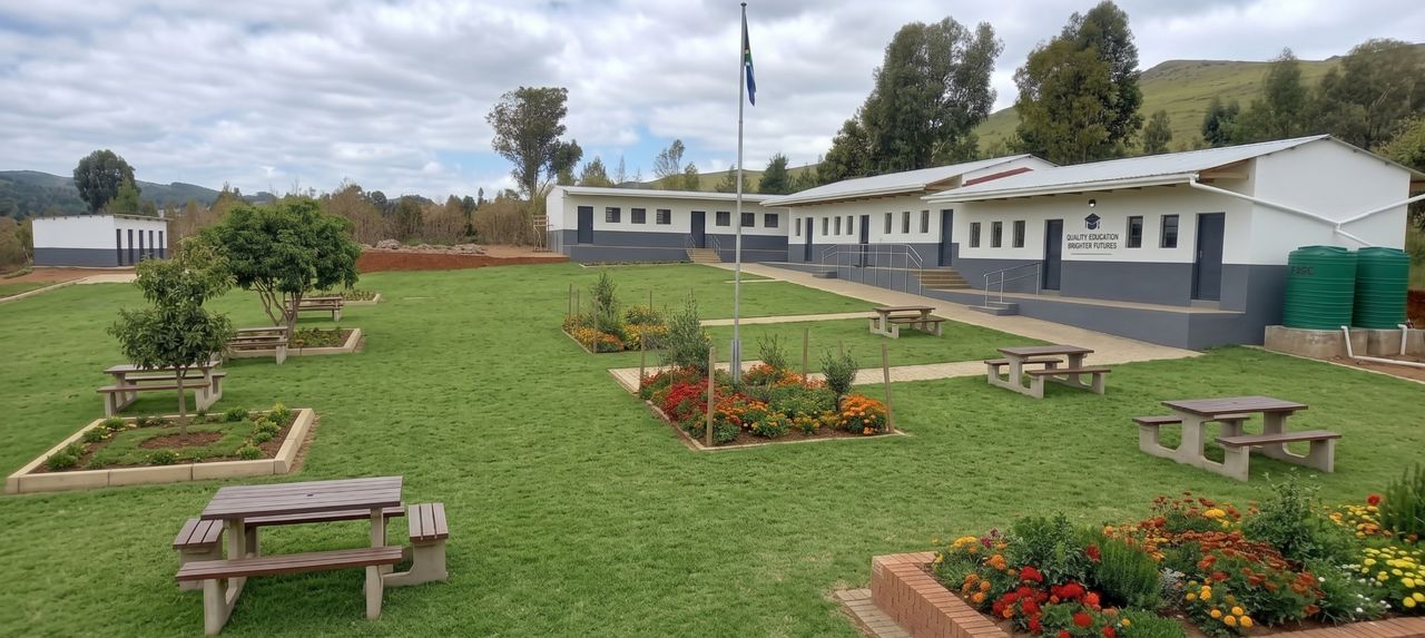Victory Primary School: white and grey classroom blocks, the flagpole, lawns with picnic tables and flower beds, and green hills behind